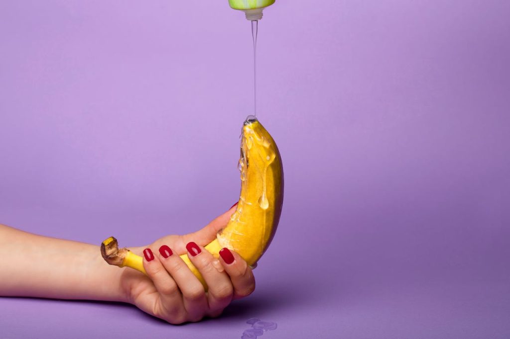 Hand holding a banana while lubricant is being poured over it against a purple background.