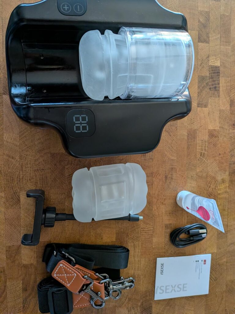 Hands Free Pocket Pussy thrusting male masturbator kit with dual TPE sleeves, wearable straps, phone holder, USB-C charging cable, lubricant, and accessories displayed on a wooden table.