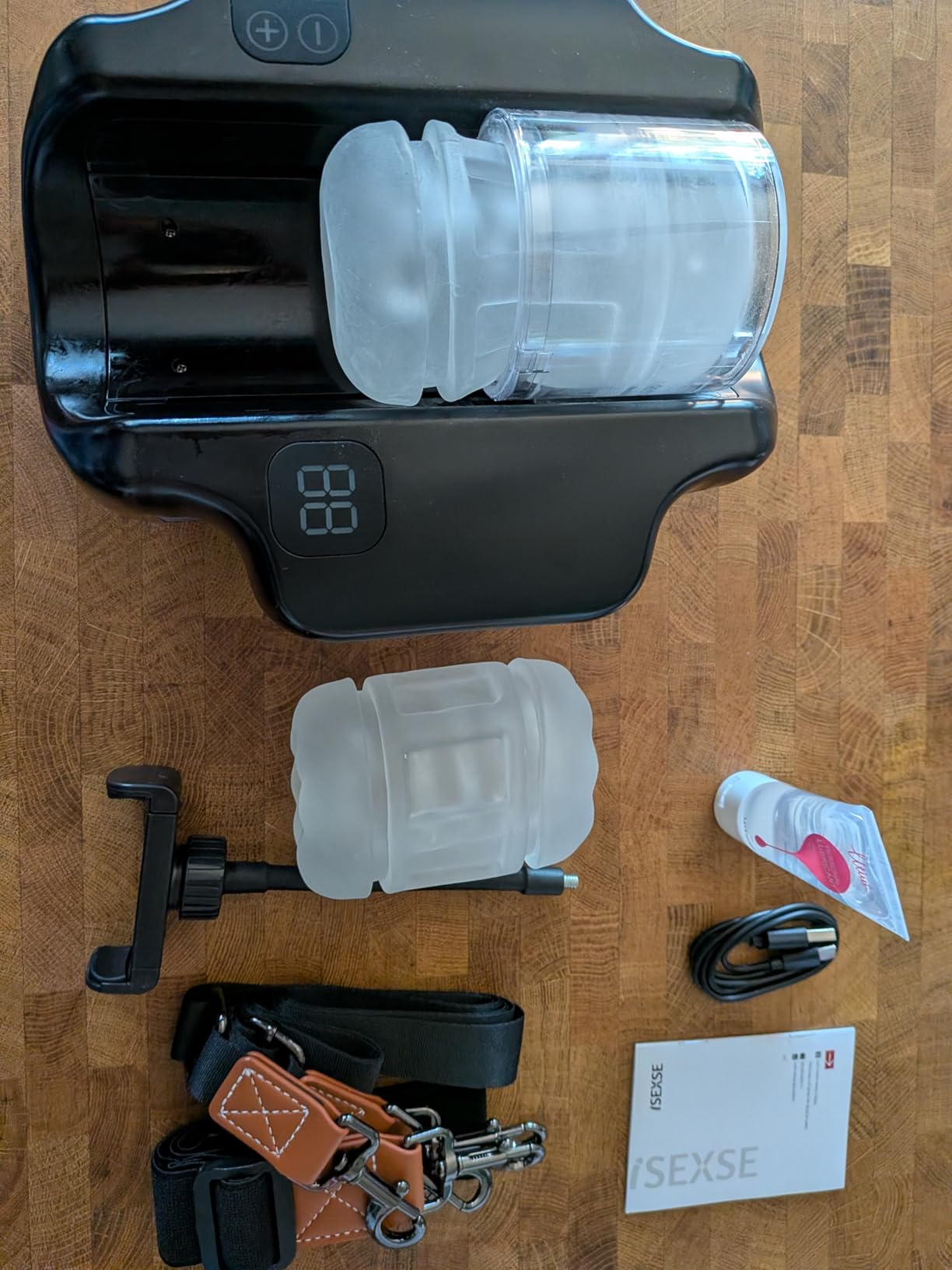 Hands Free Pocket Pussy thrusting male masturbator kit with dual TPE sleeves, wearable straps, phone holder, USB-C charging cable, lubricant, and accessories displayed on a wooden table.
